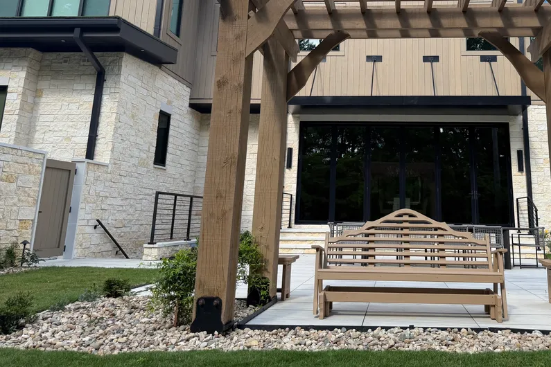 River rock bed with shrubs beside a stone-faced building, a wooden pergola and a bench on a patio