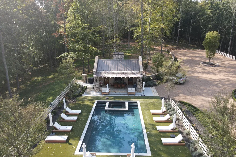 Aerial view of a backyard pool and pool house with planted beds along the fence and a gravel parking court