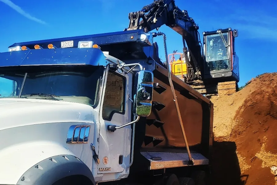 Excavator loading dirt into a tri-axle dump truck beside a cut bank on a dirt job